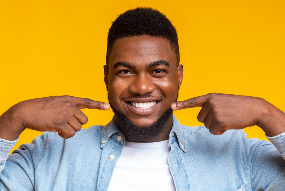 Dark-haired man with good oral health smiles and points to his healthy smile against a yellow wall